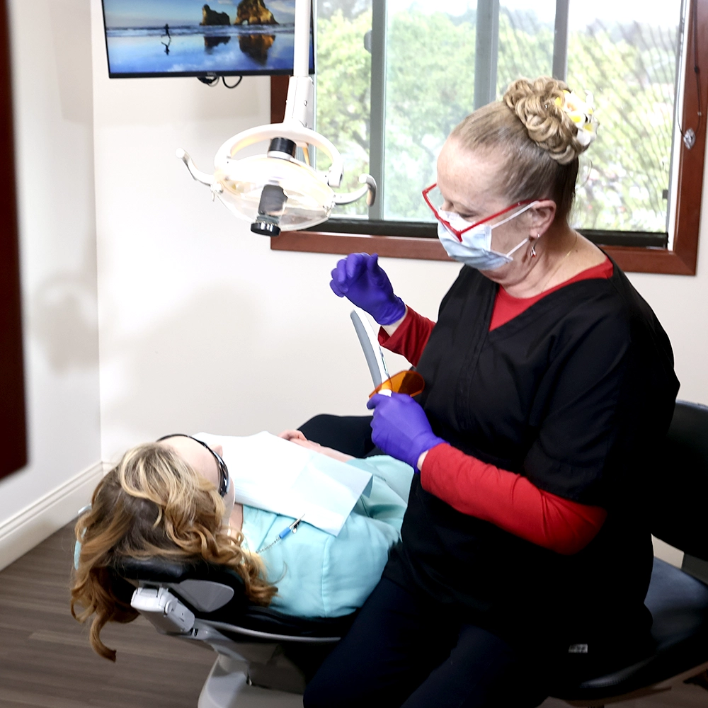 A female dentist, dressed in a black and red scrub top, working on a reclined patient wearing a turquoise dental bib. The room has a serene painting of a beachscape, adding a calming atmosphere to the dental office.