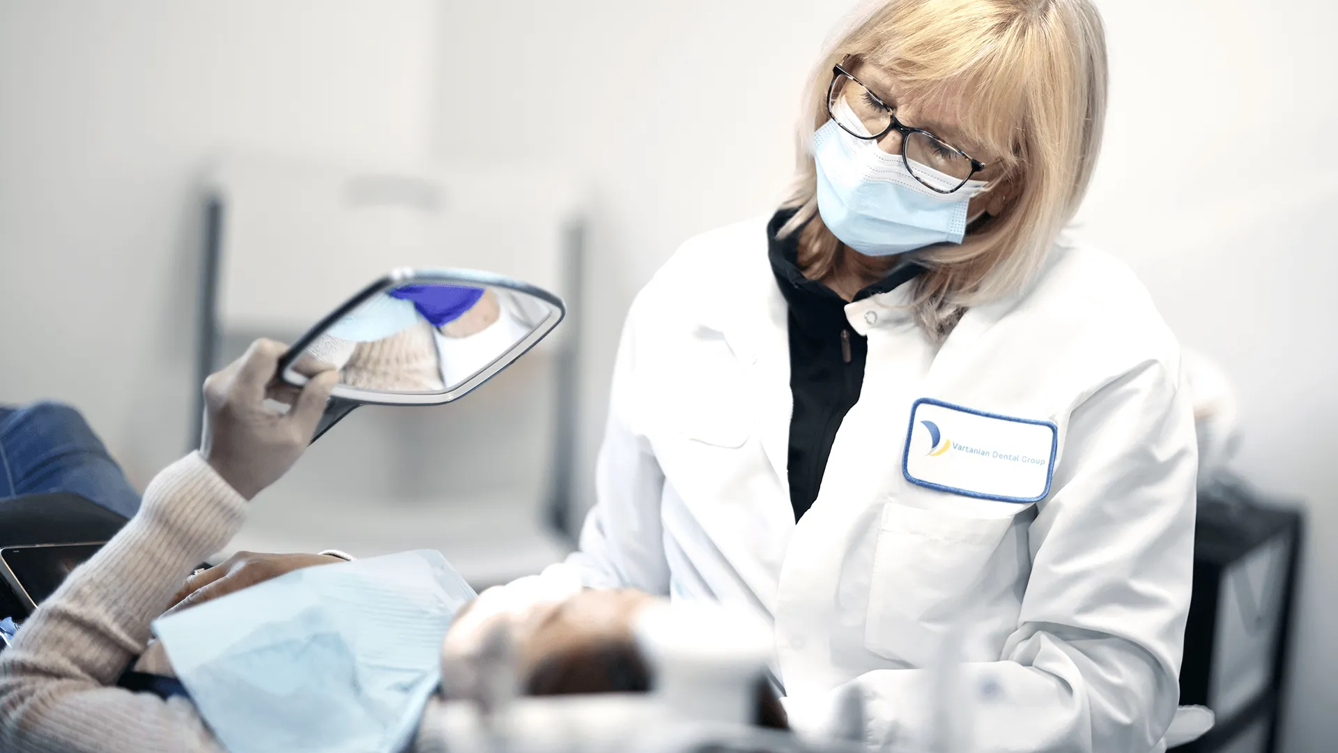 A female dentist in a white lab coat and blue mask examines a patient using a hand mirror in a well-lit dental office. The patient is reflected in the mirror, indicating a detailed dental check-up focused on dental health and patient interaction.