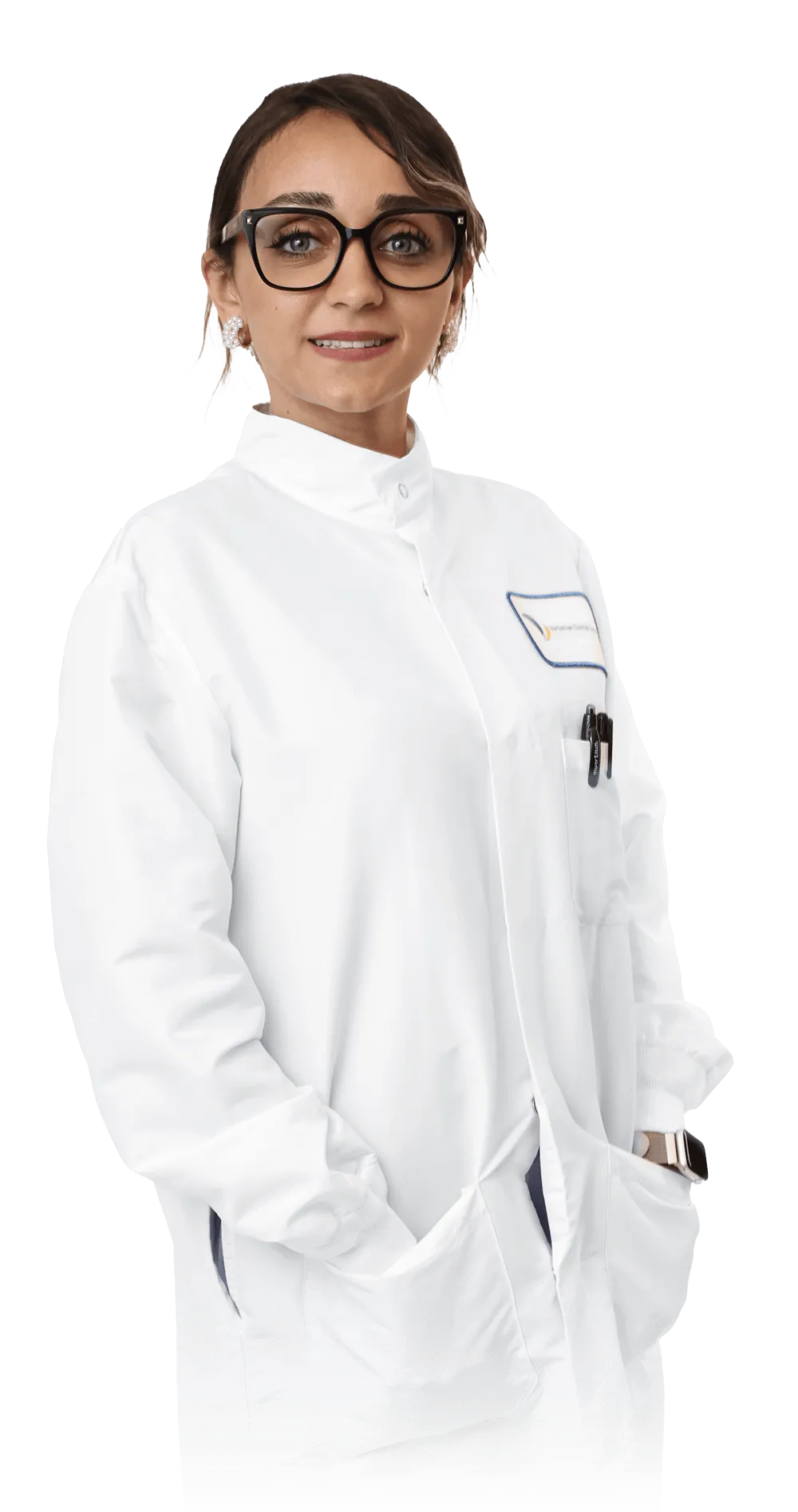 A professional portrait of Asma E., wearing a white lab coat with the Vartanian Dental Group logo on the chest pocket. She has short dark hair, wears black-framed glasses, and is smiling confidently. The dentist is holding her hands in her pockets, exuding a warm and approachable demeanor. 