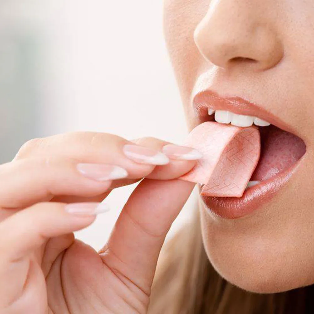 A close-up of a woman placing a piece of chewing gum into her mouth, with focus on her lips, teeth, and hand.