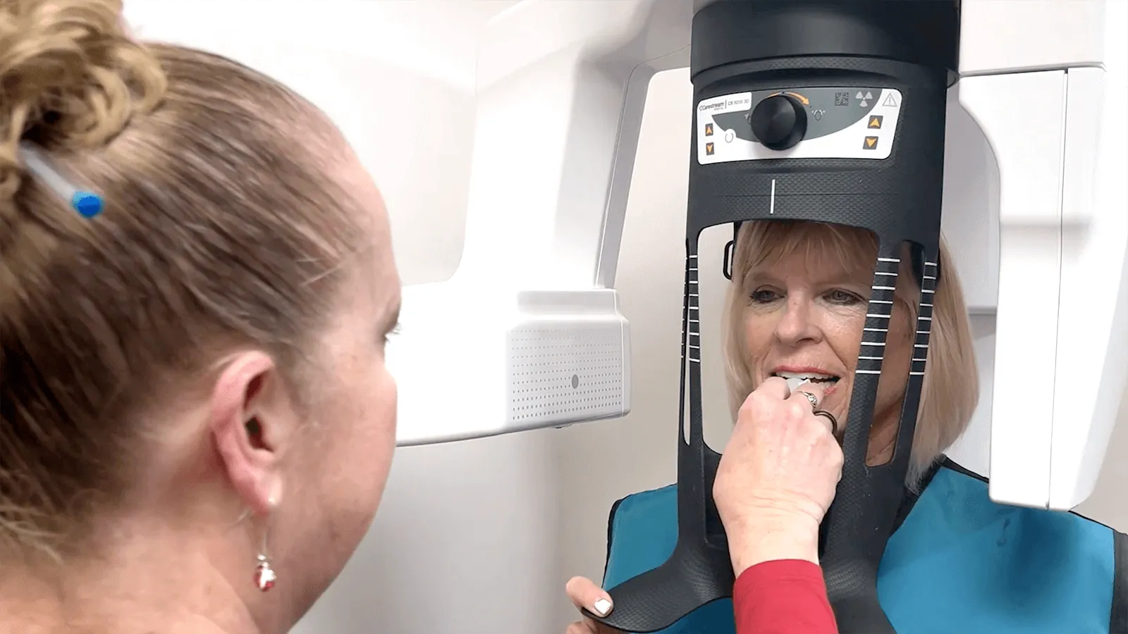 Dental assistant sitting in front of a screen displaying dental X-rays while the dentist works in the background.