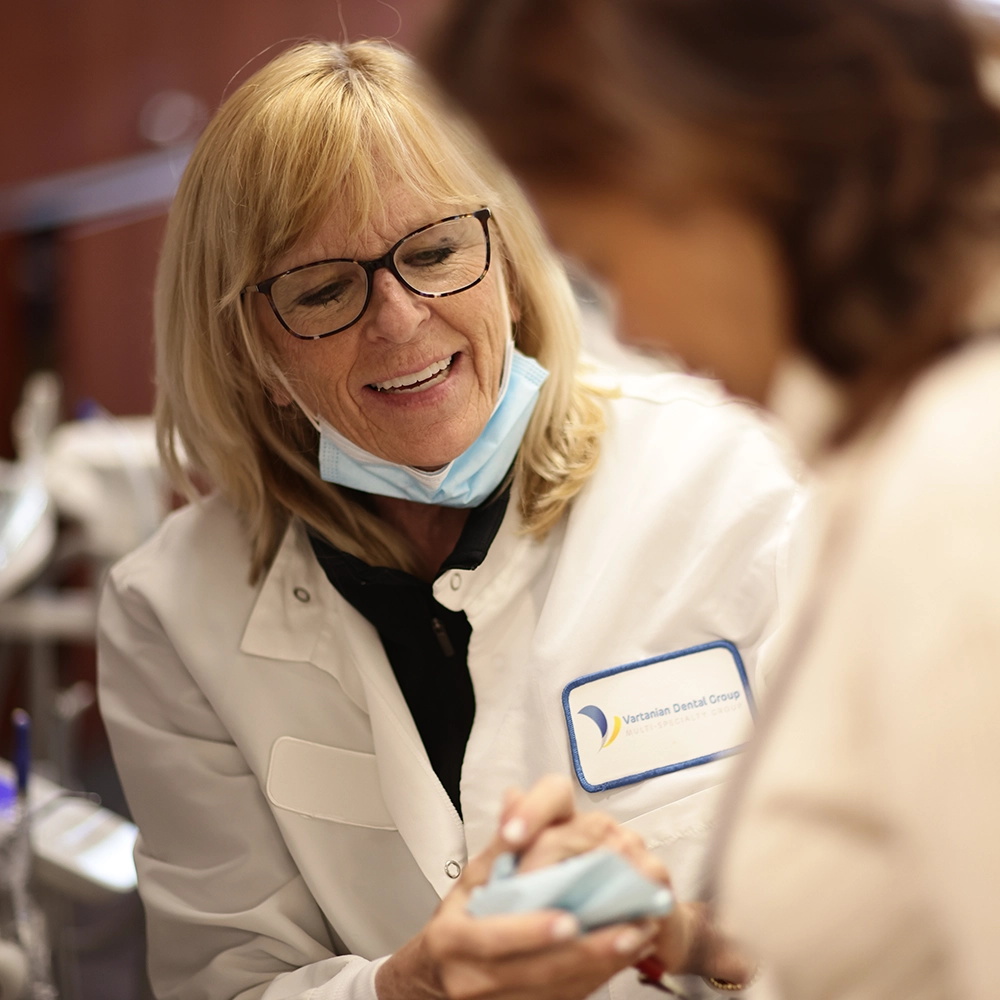 A female dentist wearing a white lab coat and blue surgical mask demonstrates a dental tool or model to a patient in a warmly lit dental office.