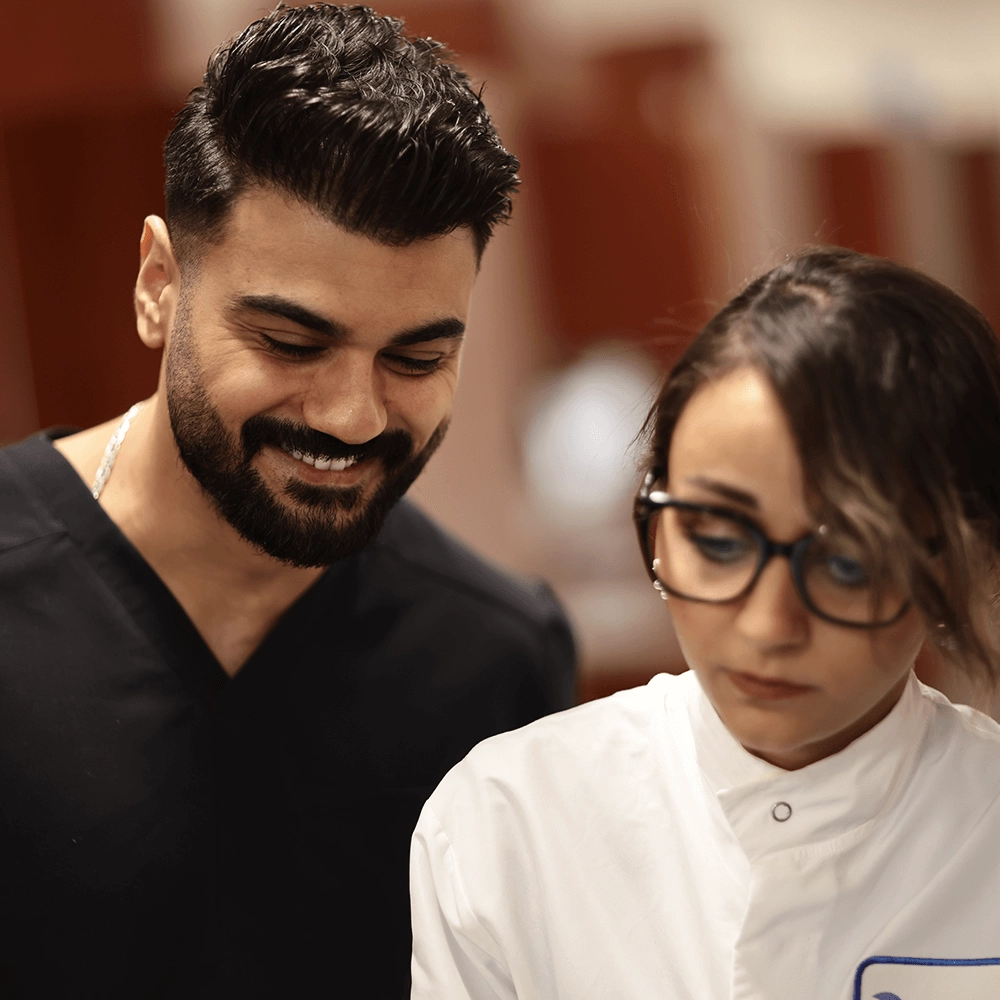 A male dentist and a female dental assistant, both in black scrubs, focus intently on a dental model. 