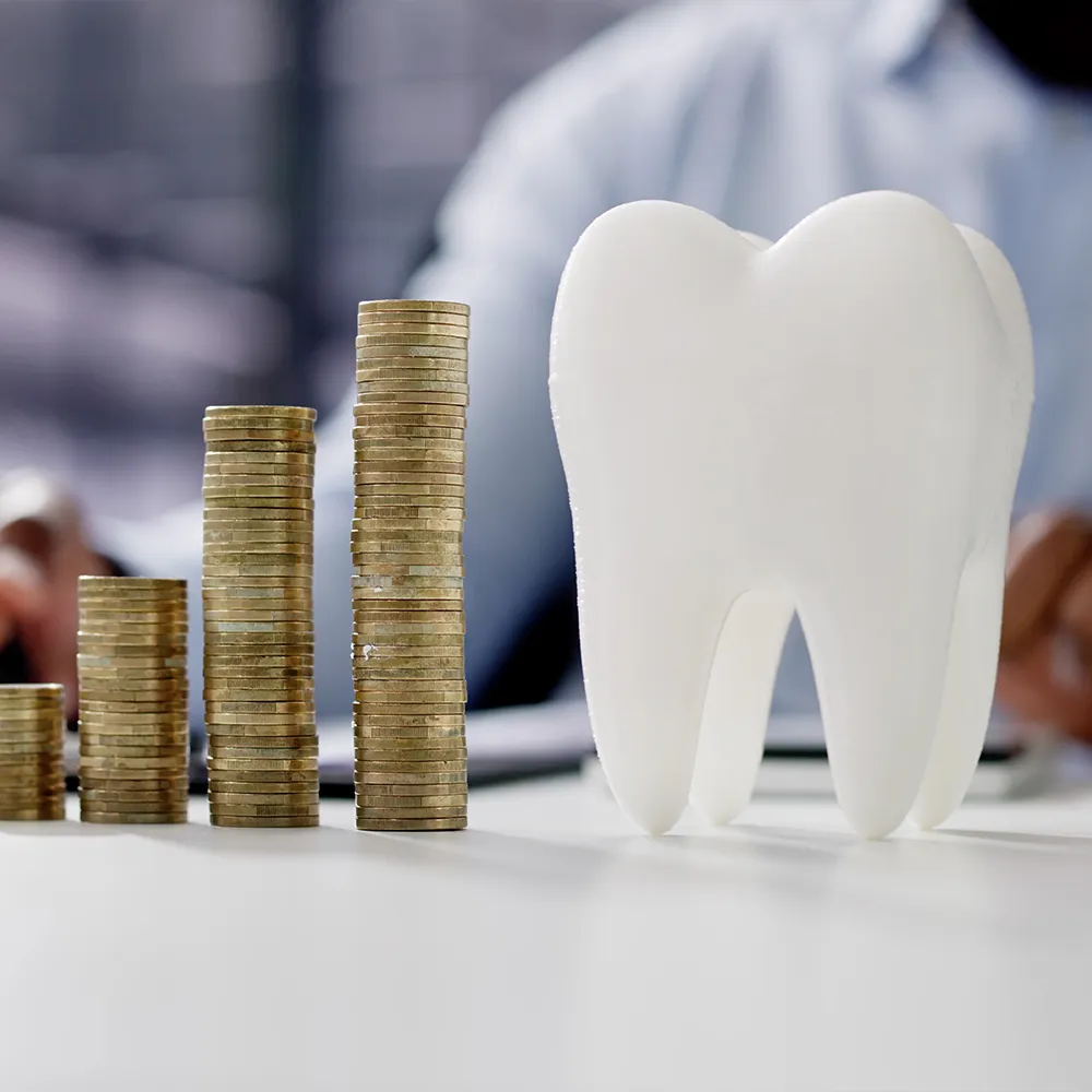 A conceptual image featuring a large white tooth model beside ascending stacks of coins, symbolizing the cost of dental care. In the blurred background, a person in business attire is partially visible, representing financial planning and investment in dental health.