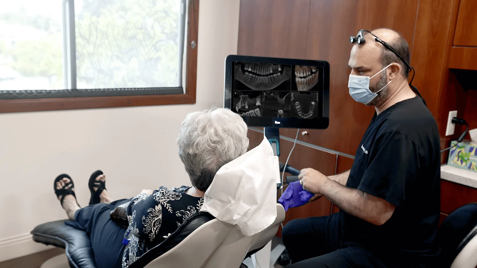 An elderly woman seated in a dental chair, looking at dental x-rays displayed on a monitor while the dentist, Dr. James Vartanian, wearing a surgical cap, glasses, and a mask, observes and discusses the findings.