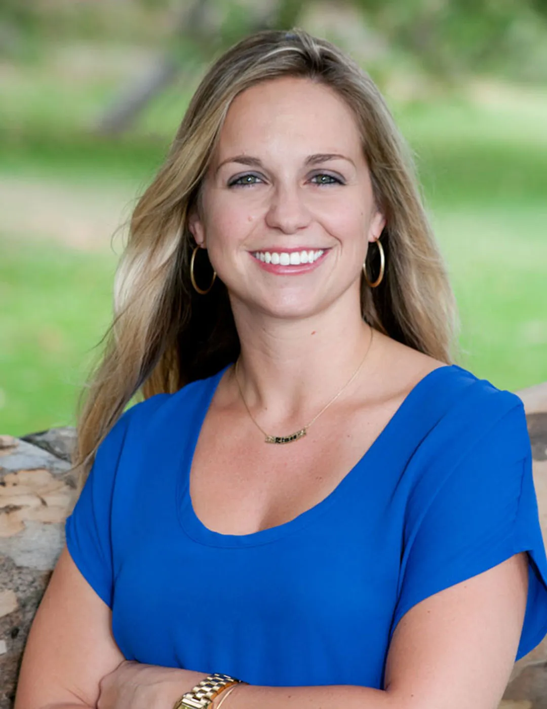 A portrait of Dr. Aimee Trujillo, wearing a vibrant blue top and a delicate necklace, smiling broadly in a natural outdoor setting.