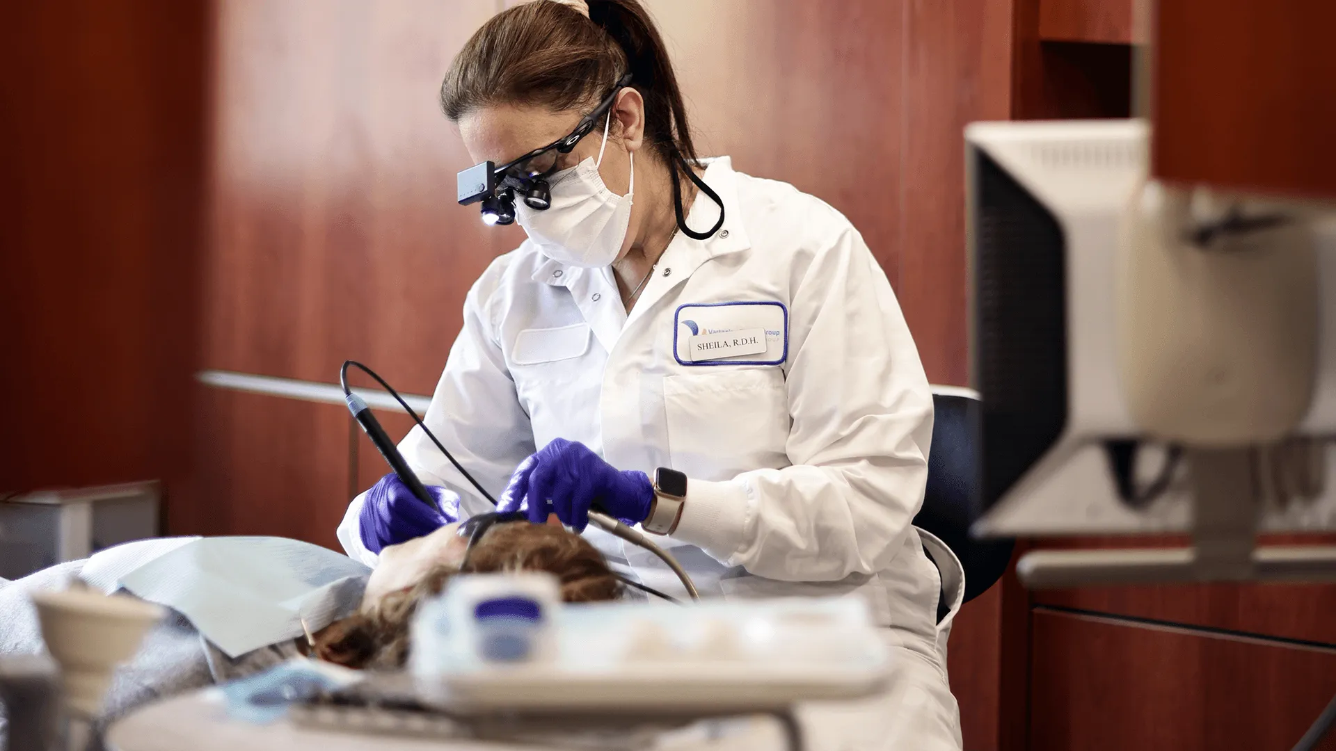 A dental hygienist focused on cleaning a patient's teeth in a modern dental office. She wears protective glasses, a mask, and gloves, employing advanced dental tools and a computer monitor to guide her work.