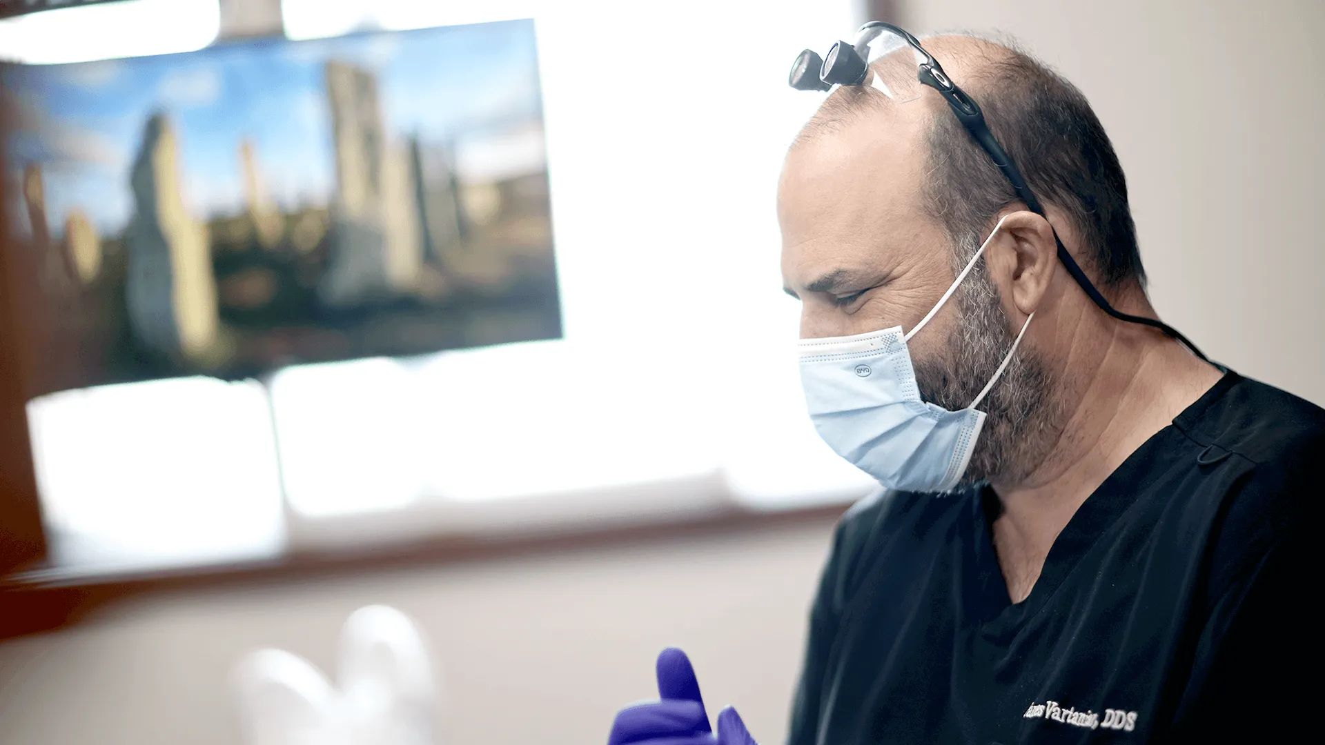 Focused side profile of Dr. James Vartanian, a bald, middle-aged male dentist wearing a black scrub, surgical mask, and purple gloves.