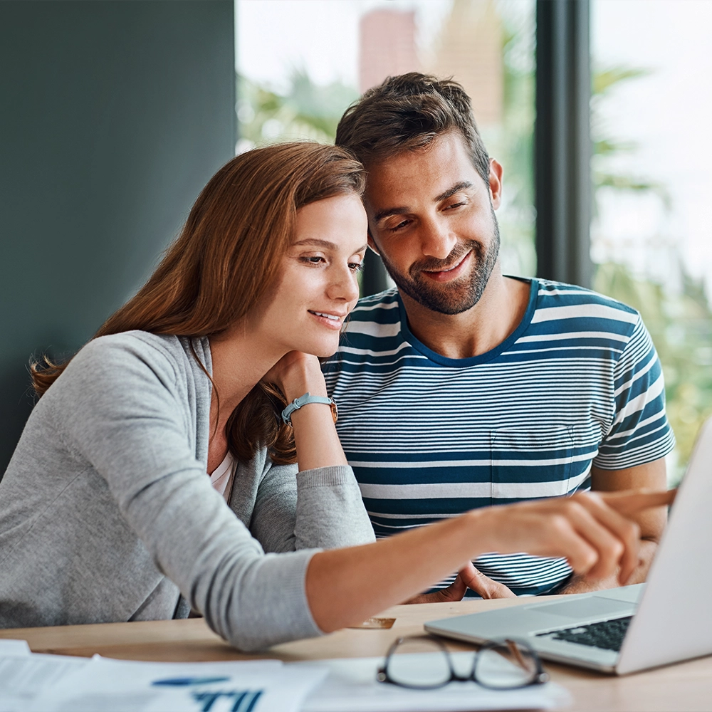 A smiling woman and man closely view content on a laptop together, possibly evaluating dental insurance options. The woman has long red hair, and the man has short brown hair and a beard, wearing a striped blue and white T-shirt.