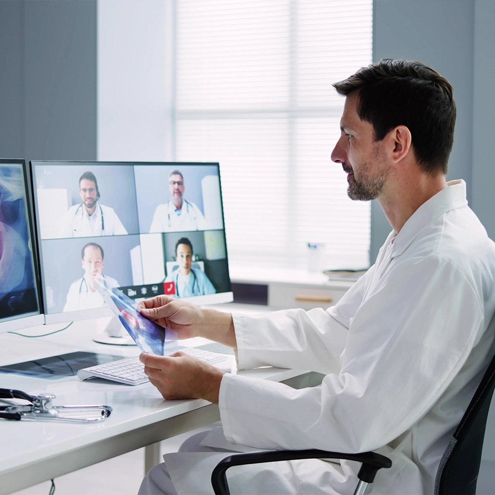 Male dentist in a white lab coat reviews a dental X-ray while participating in a virtual meeting with multiple colleagues on computer screens, showcasing a blend of technology and healthcare.