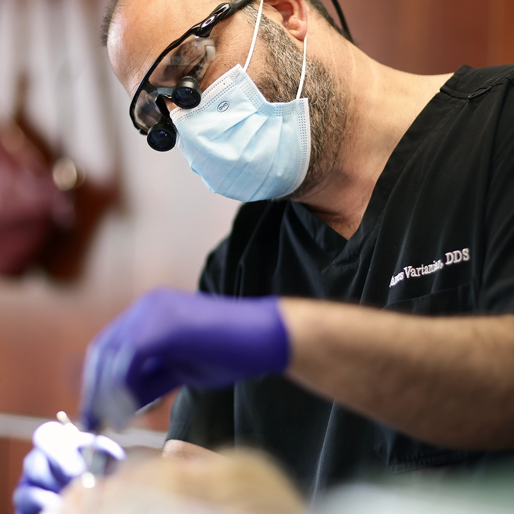 Dr. Vartanian, wearing a mask and magnifying glasses, carefully performing a dental procedure on a patient.