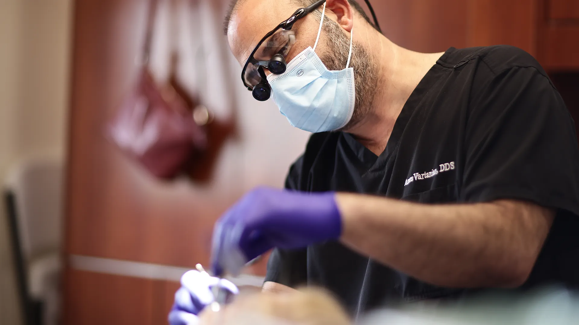 A focused male dentist, wearing a black scrub top and a surgical mask, working on a patient in a dental clinic. He is wearing gloves and a magnifying loupes on his eyes.