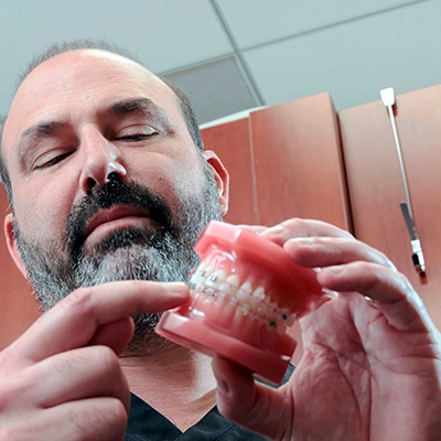 A male dentist, holding a dental model, explains orthodontic appliances to the viewer, demonstrating an educational moment in a dental office.