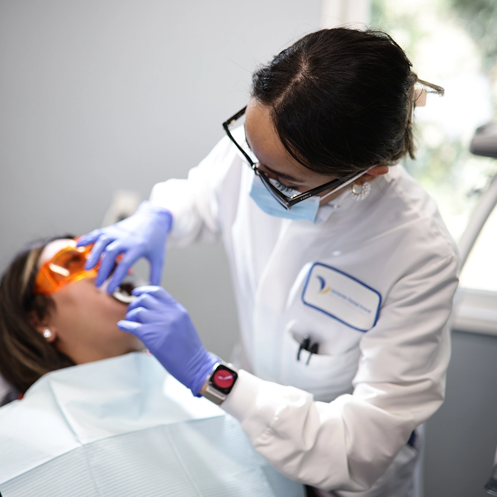 A female dentist in a white lab coat and protective eyewear is carefully applying a teeth whitening gel to a patient’s teeth. The patient is seated in a dental chair, wearing orange protective glasses, ensuring safety during the procedure.