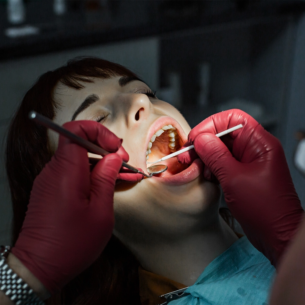 A dental patient undergoing a procedure with her mouth open, showing dental equipment in use. The dentist, wearing red gloves, performs the procedure in a well-equipped dental clinic, highlighting the comprehensive care provided under dental insurance coverage.