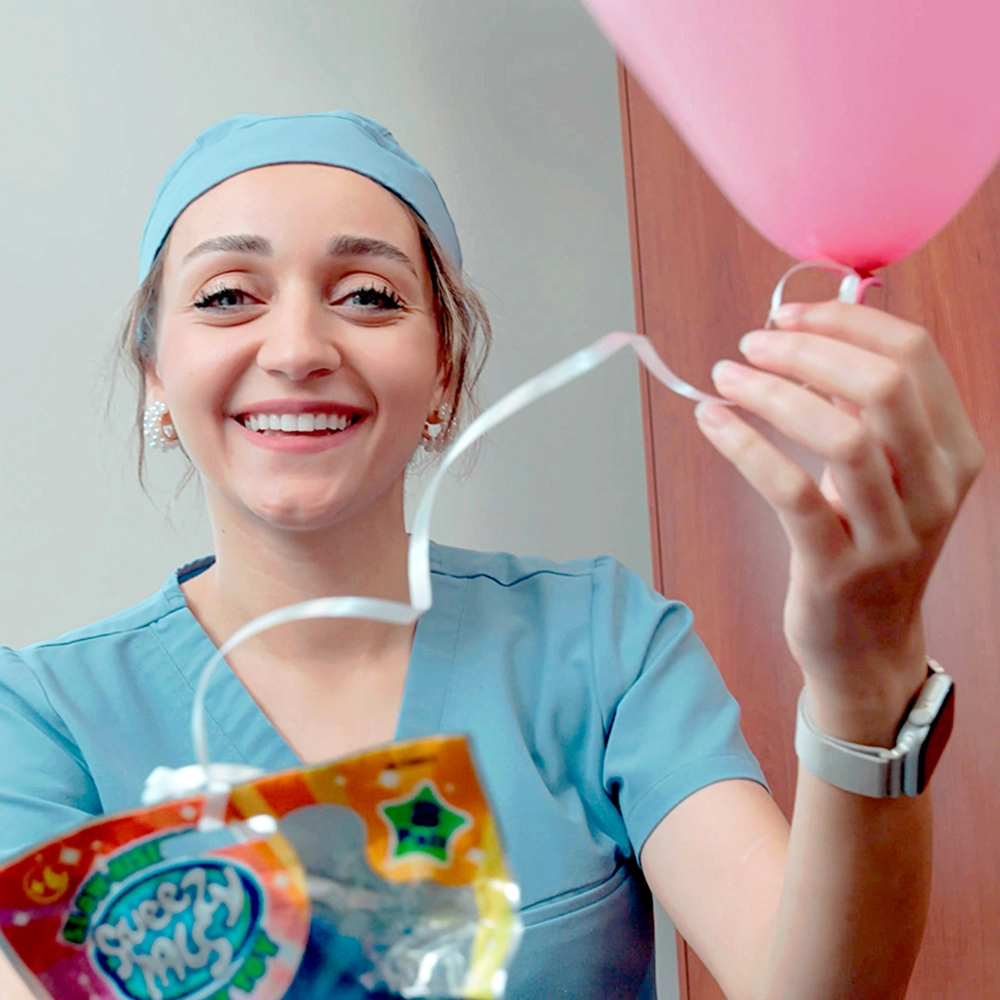 A cheerful dental assistant in a turquoise scrub cap and matching scrubs holds a pink balloon and a package of children's dental products.